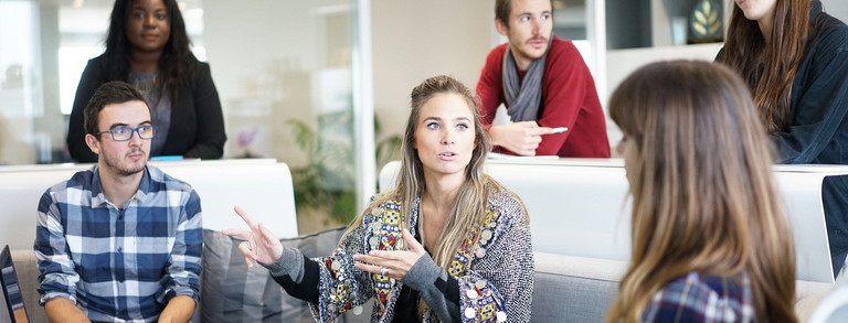 sechs Erwachsene sitzen in einem Büro und diskutieren miteinander, während sie Unterlagen und Computer bereit gelegt haben.
